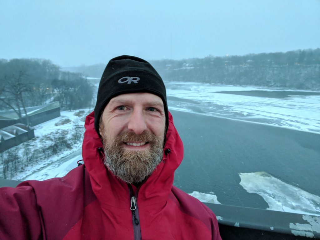 Bearded man with black stocking cap and red winter jacket with gray river and gorge in background