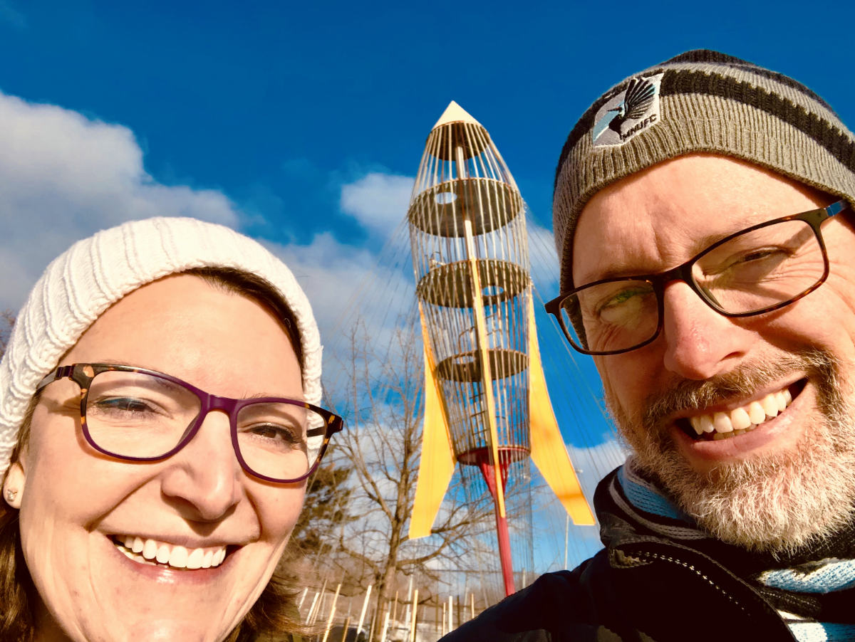woman's and man's faces closeup wearing stocking caps with yellow and red rocket sculpture in background against blue sky with clouds