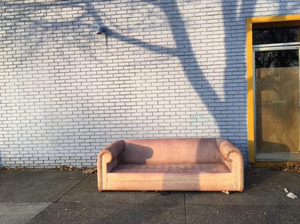 tan leather couch on sidewalk in front of white brick wall with tree shadow