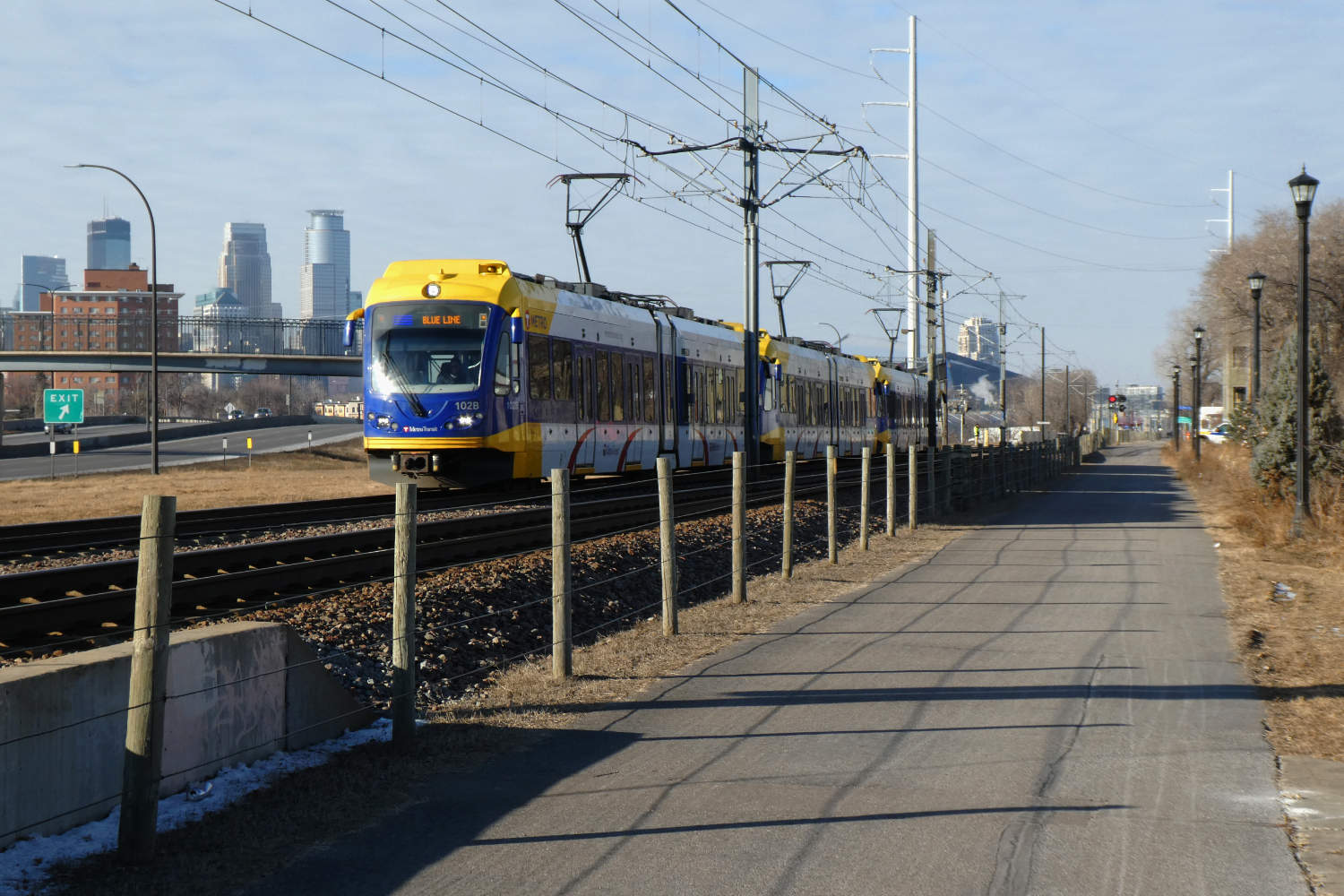 Approaching light rail train with bicycle path and fence foreground and downtown Minneapolis skyline background on a sunny day