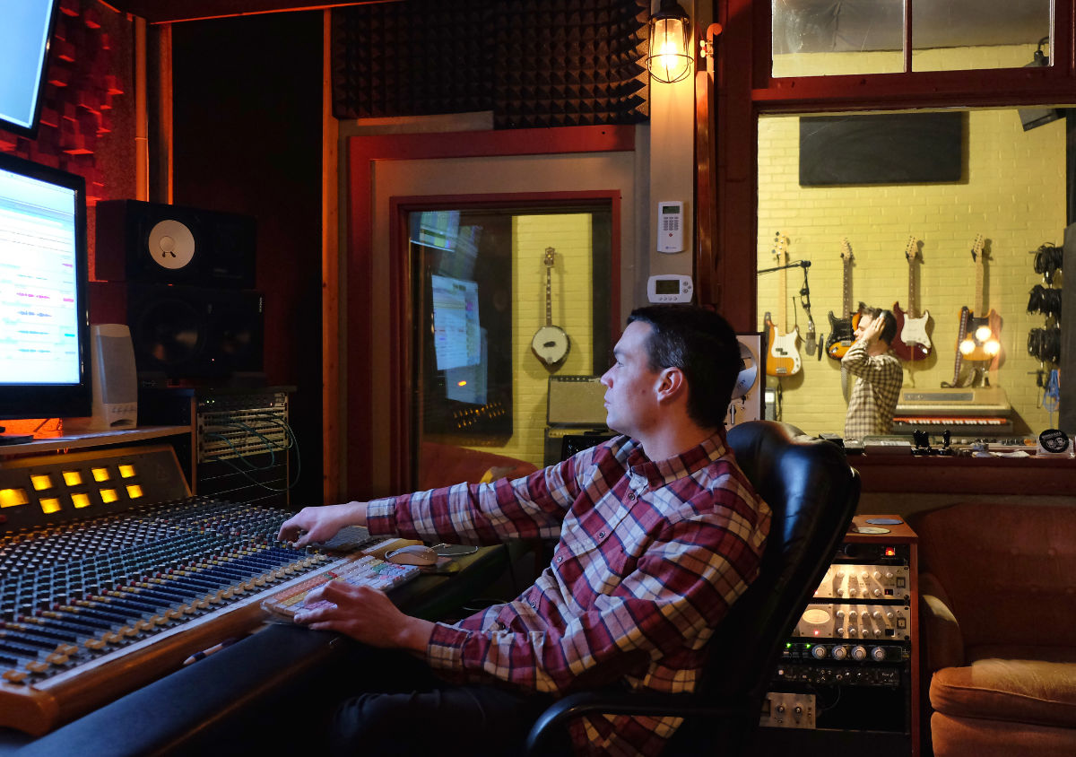 man at control board with many knobs facing monitors with background window of man with many guitars