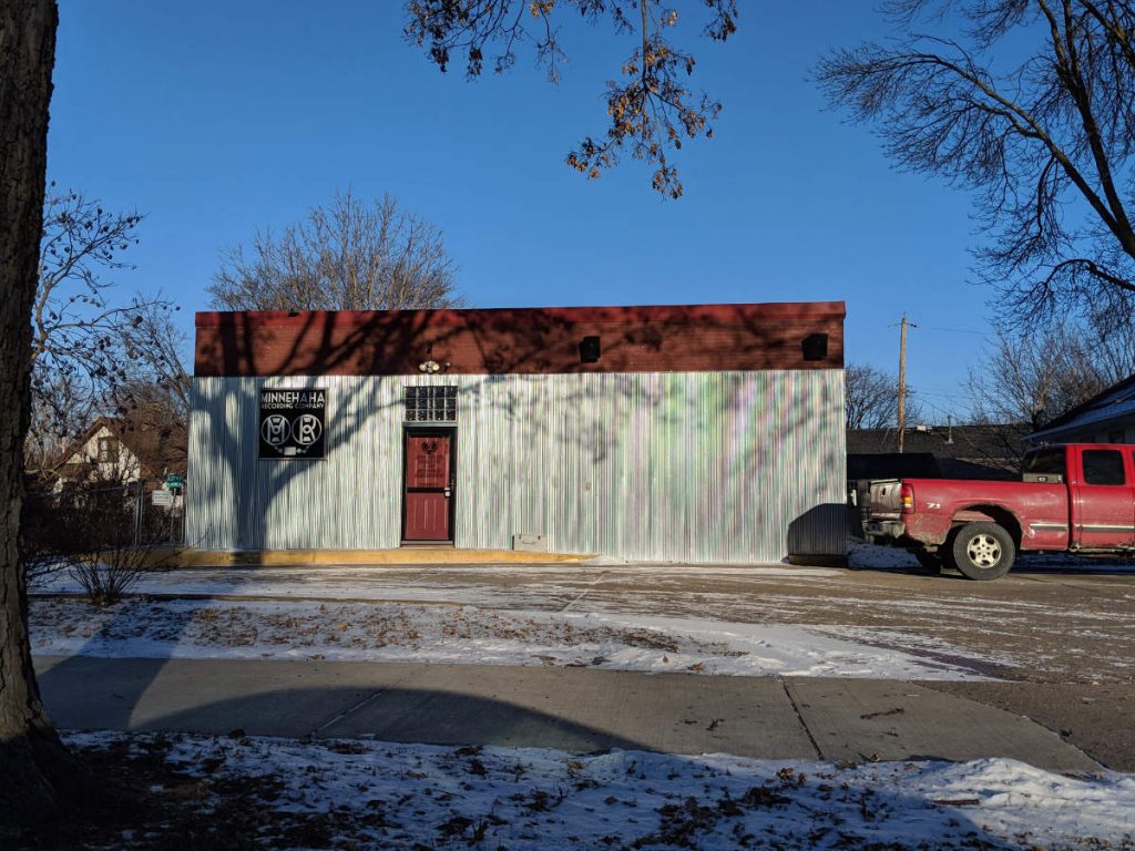 small commercial building with iridescent white wall, shadowed by tree on left, next to red pickup truck on right with blue sky background and street with snowy cement foreground