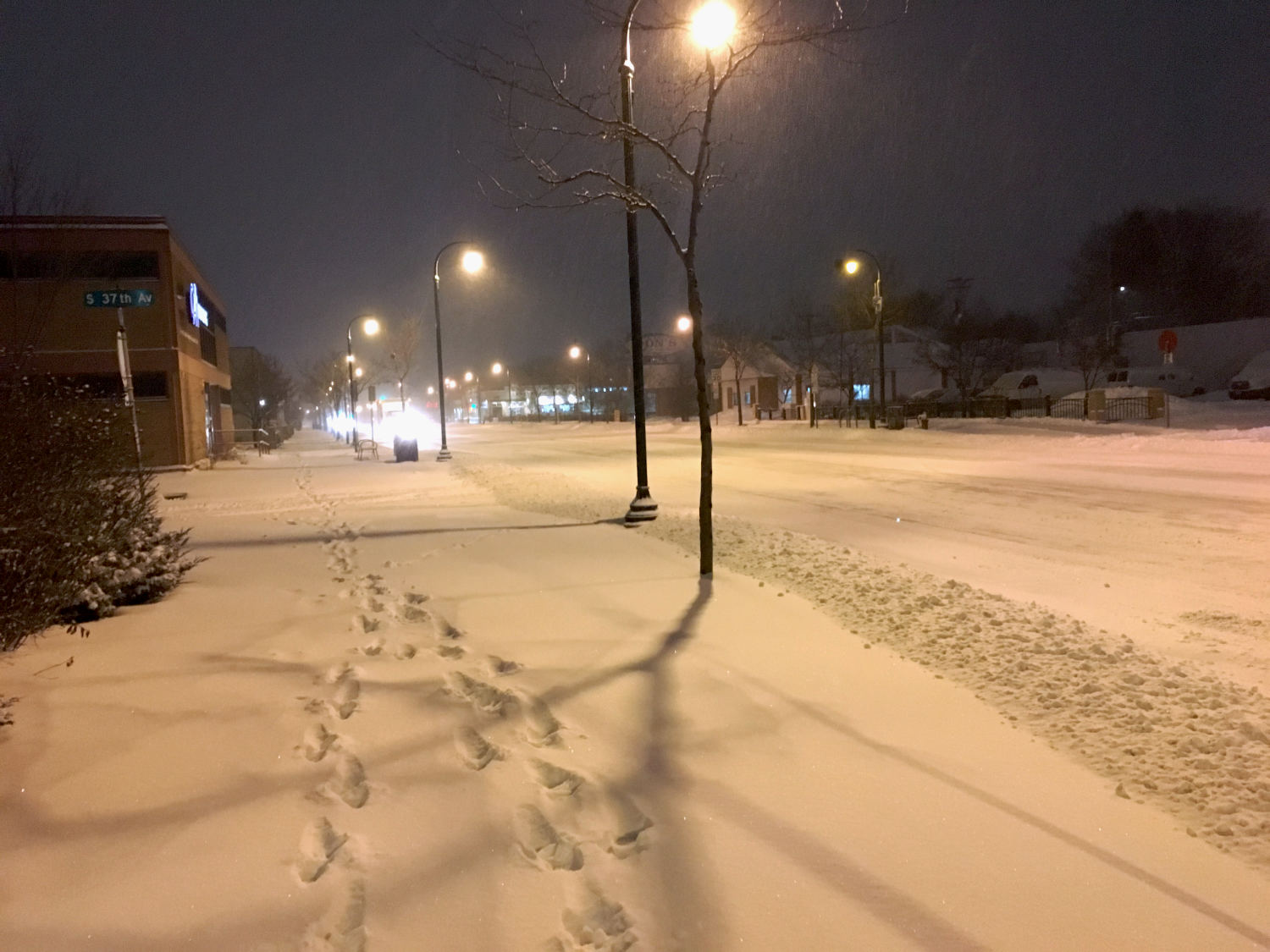 footsteps through snow-covered sidewalk with snowy street under streetlights in the dark