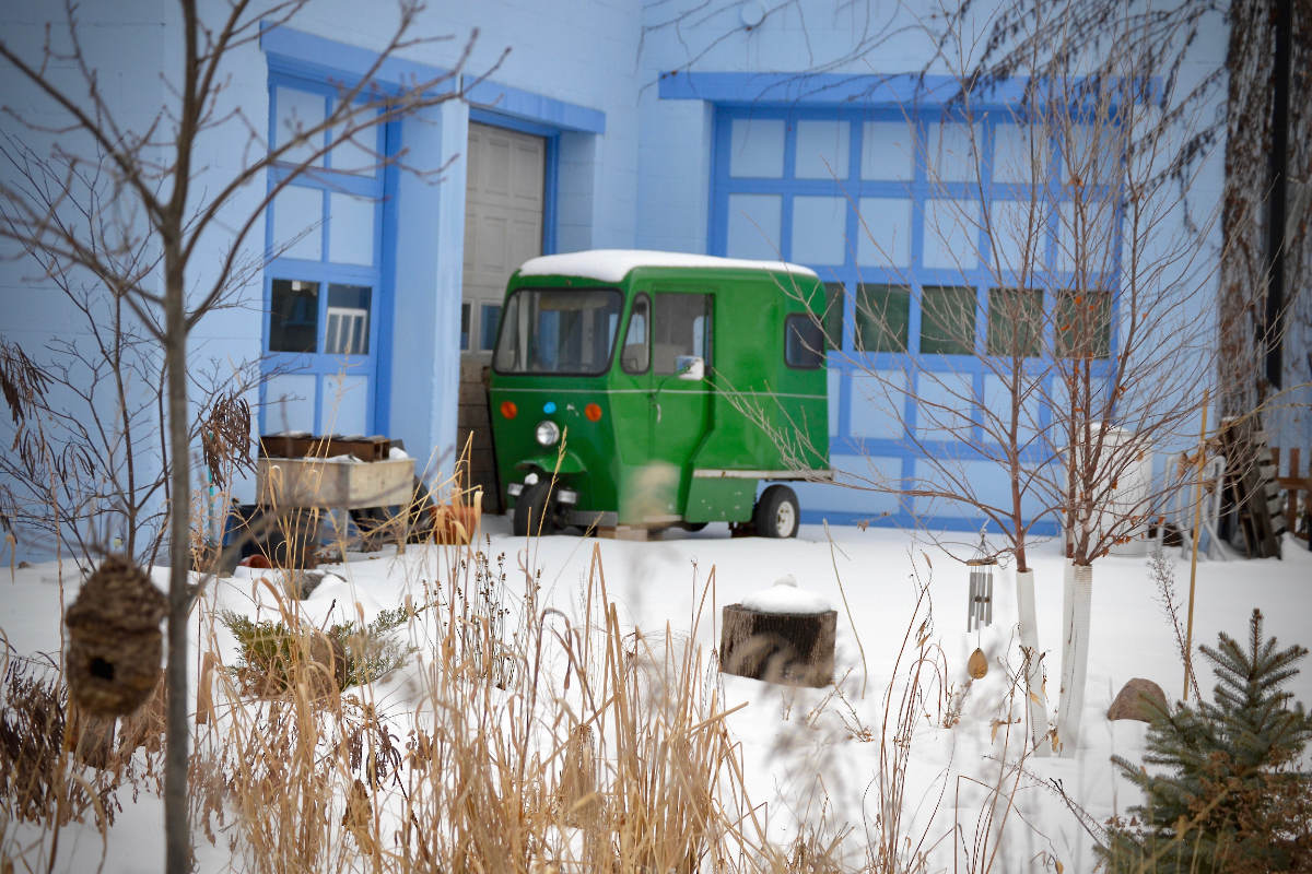 Small green mini-van parked by lue building with snow and tall dried grasses in foreground