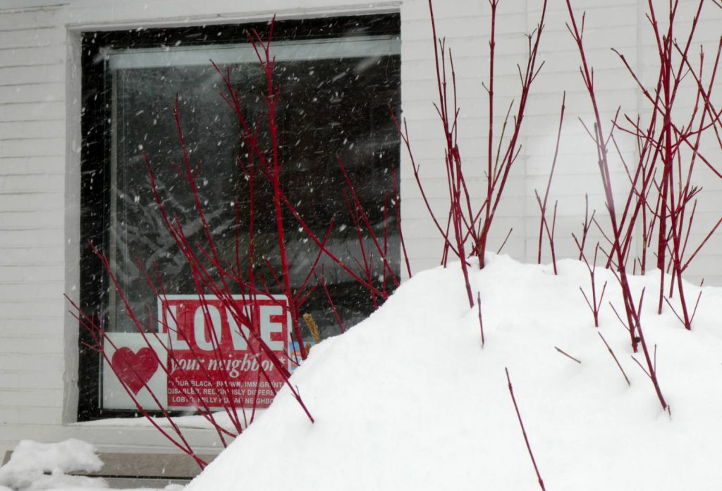 red signs in window, one a red heart, another reads Love Your Neighbor, with red branches in a snow mound in foreground