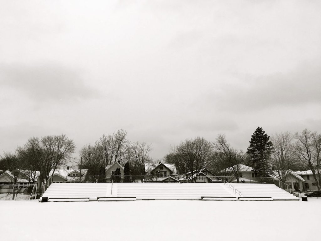 long set of bleachers covered in show with a snow-covered field in foreground and bungalow houses and trees in background