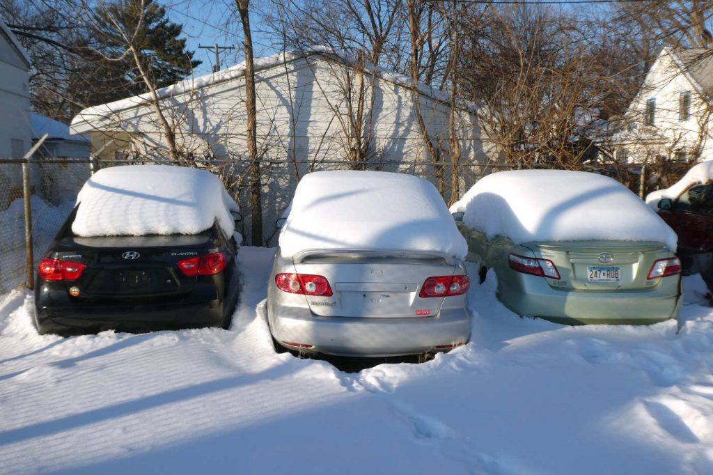 Rears of three small sedans covered in snow in a snowy lot