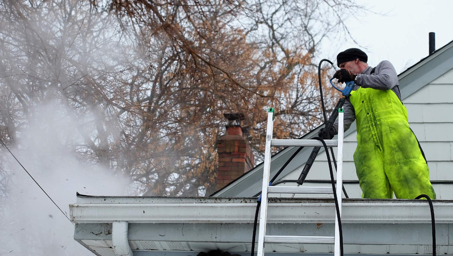 man in yellow-green safety overalls by a ladder on a roof spraying steam from a long metal tool