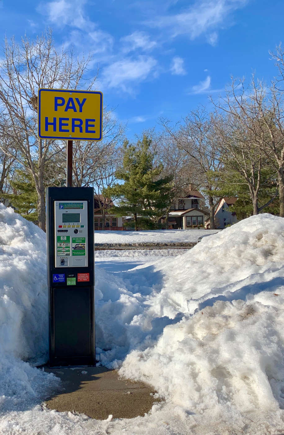 parking meter station with big Pay Here sign on top surrounded by mounds of snow on a sunny day