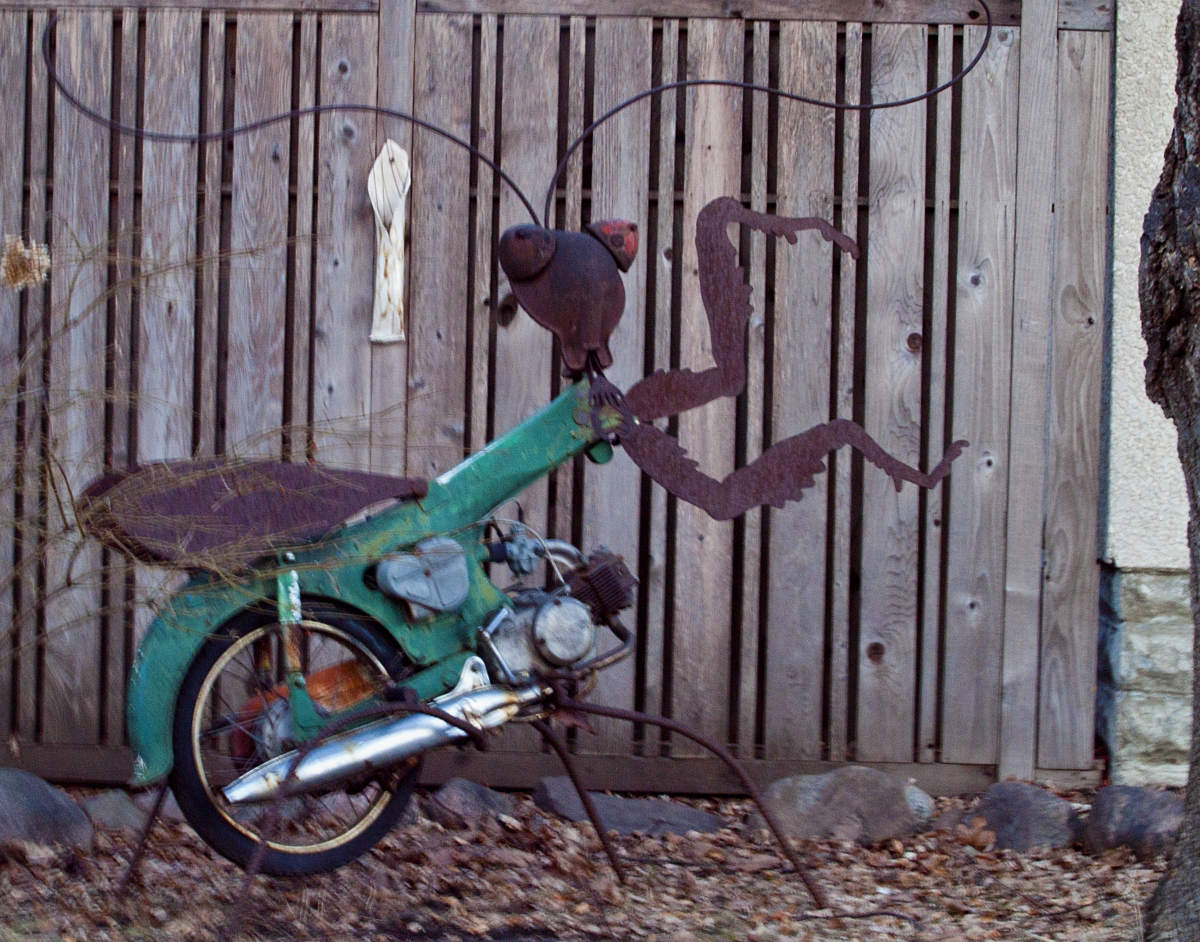 green motorcycle with rusty sculpture replacing front wheel and handlebars to look like a praying mantis