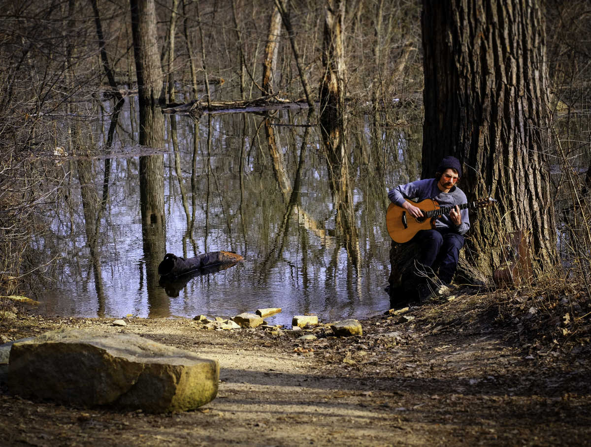 man sitting against a big tree trunk and playing guitar next to flooding waters of a creek