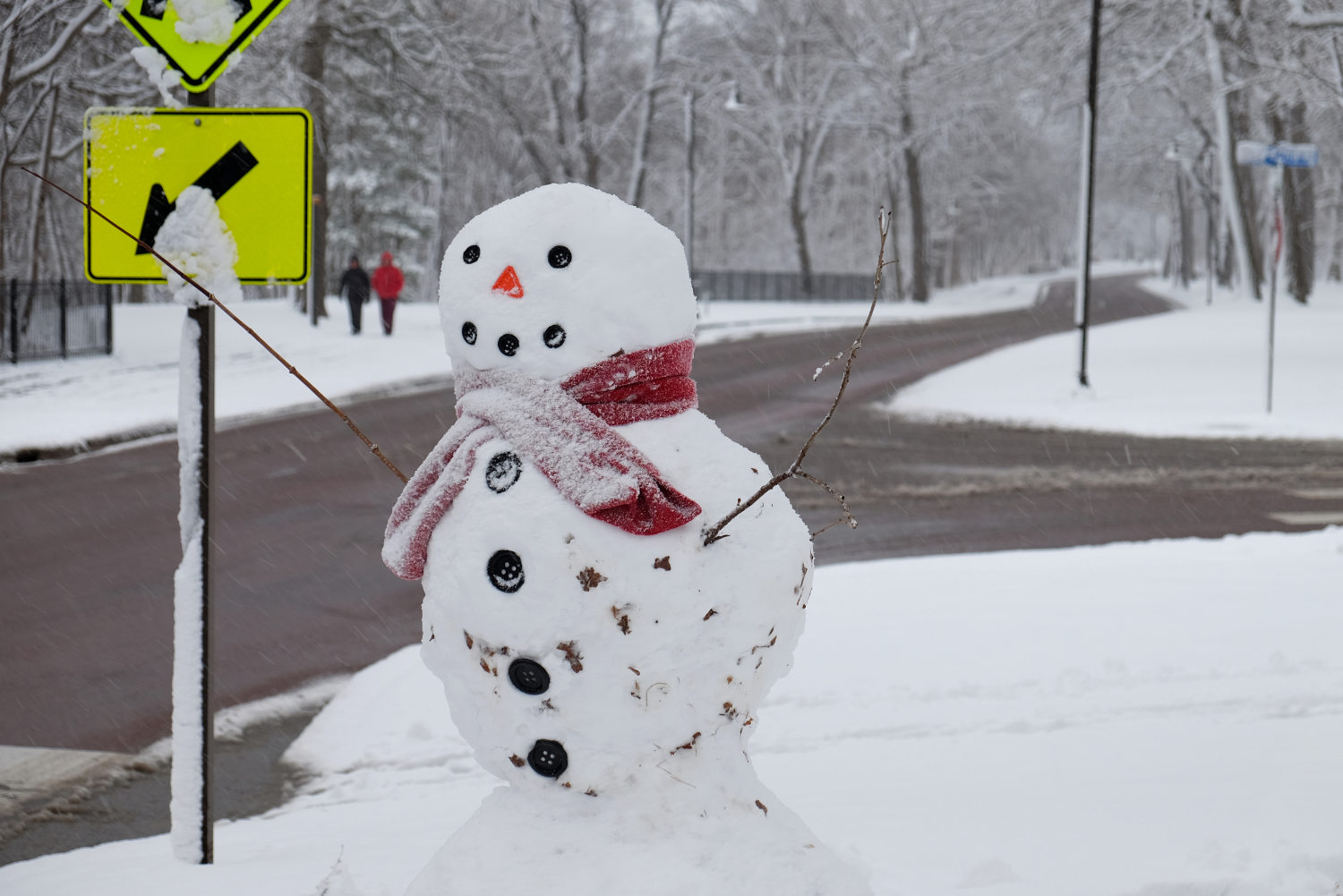 snowman detailed with black buttons and an orange triangle nose, with b yellow sign with arrow and couple walking in background