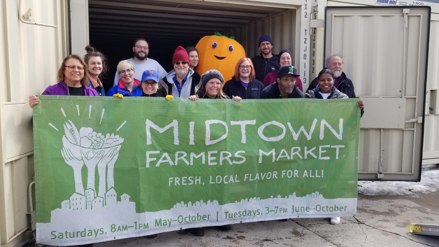 Group of fourteen people behind a large cloth sign for MIDTOWN FARMERS MARKET plus a smiley face atop a human-size orange carrot