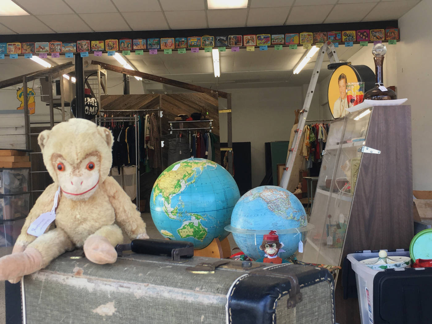 store window with globes stuffed monkey on an old suitcase surrounded by small knickknacks with racks of clothing in the background