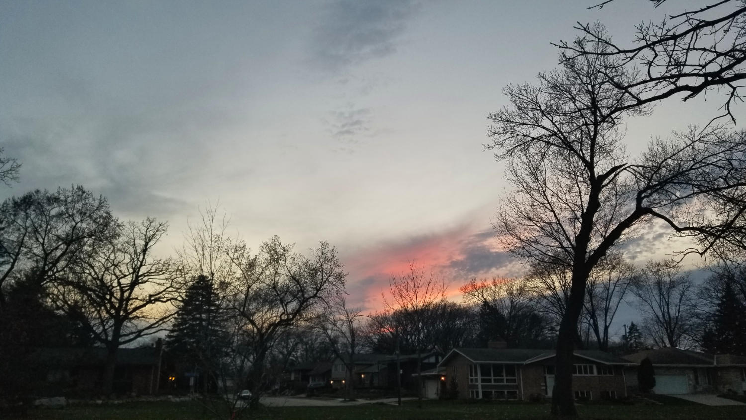 Pale orange sunset sky with clouds over dark houses and trees