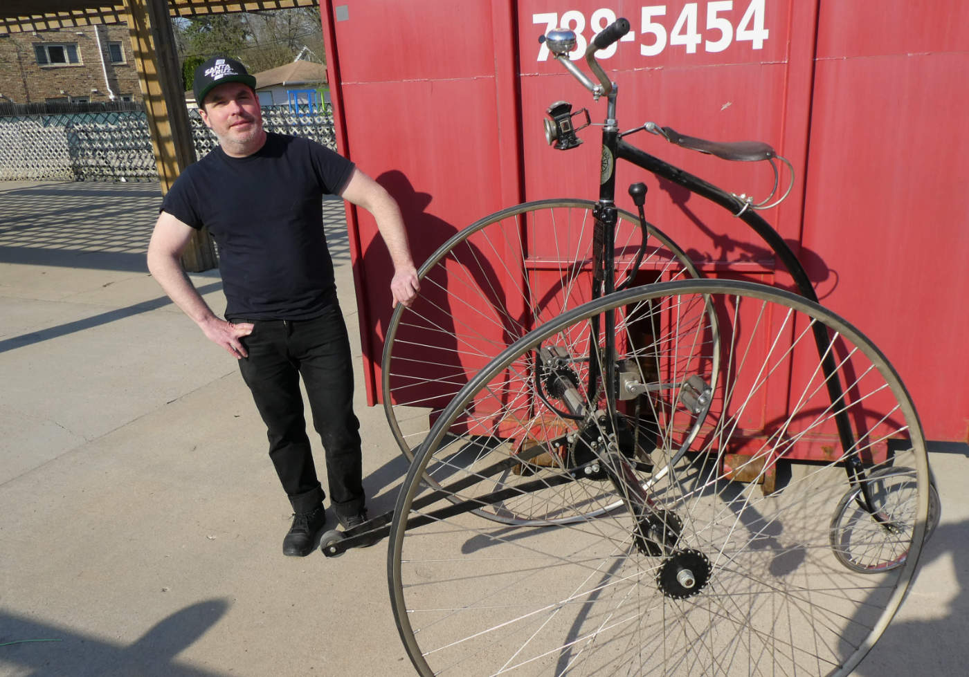 man dressed in black pants Tshirt and baseball cap standing next to a classic tall two-wheeled bicycle in front of a large red metal container box