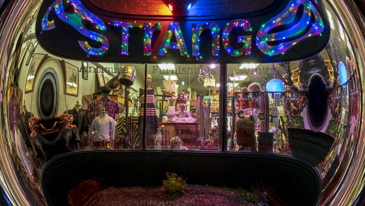 Store window filled with array of vintage items including mannequins, clothing, cactus, and a multi-colored sign reading STRANGE