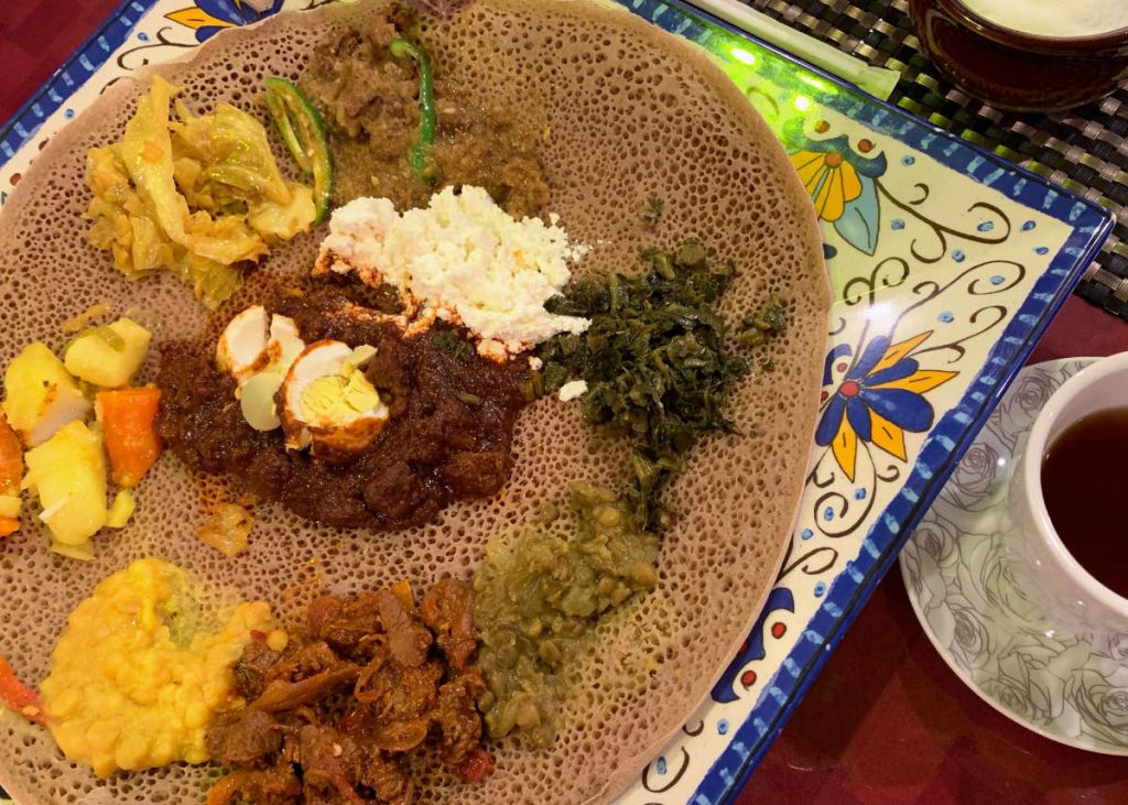 Yellow and green vegetables, brown beef and sauces, on tan bubbly-surfaced round of injera bread on a colorful square platter