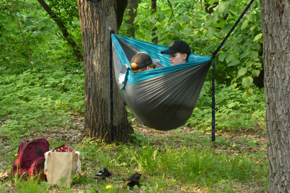 two heads with dark baseball caps sticking out of a hammock strung between two tree trunks amidst greenery