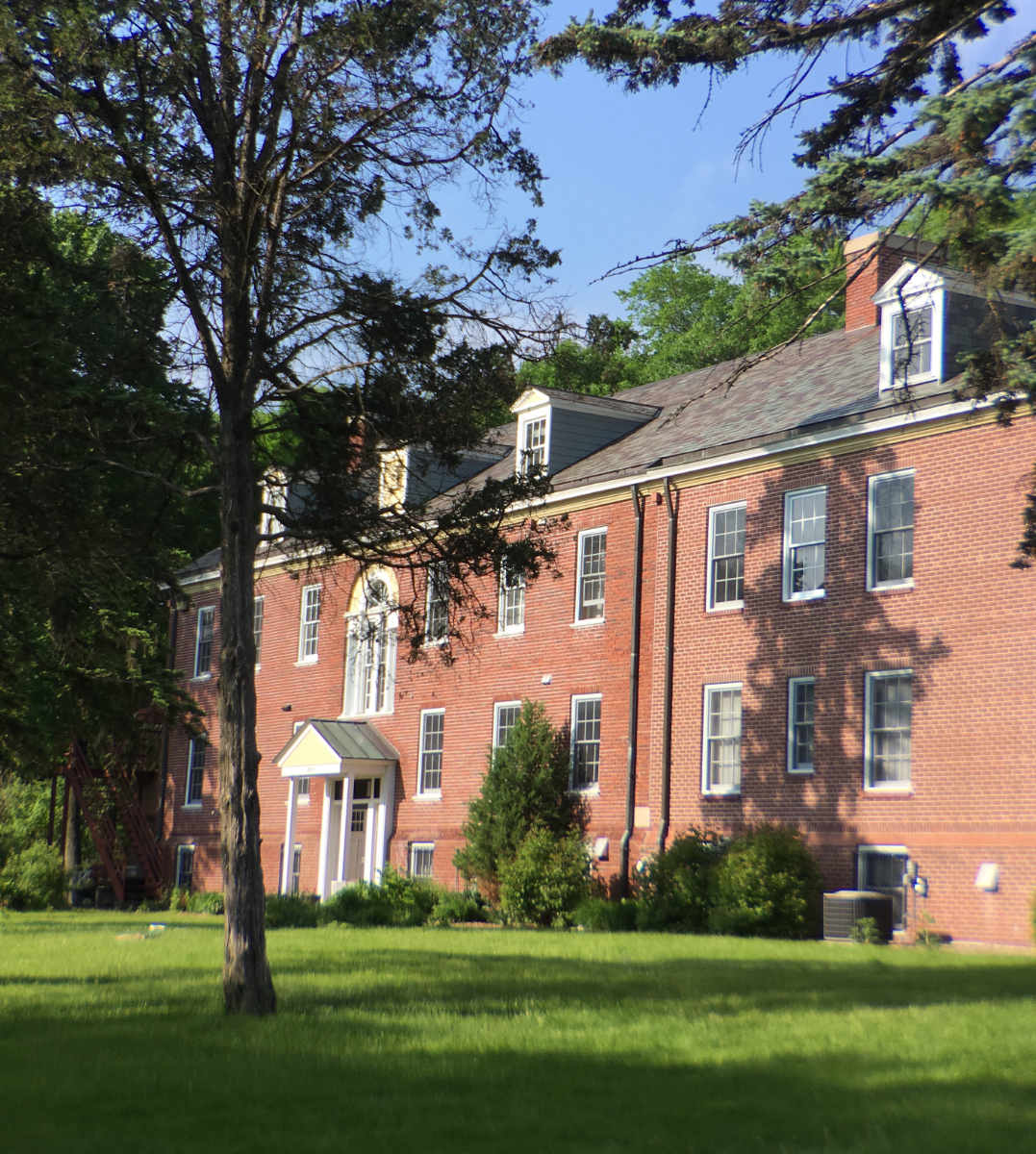 large two-sorty red brick building with dormers on the roof with a sunny lawn and trees