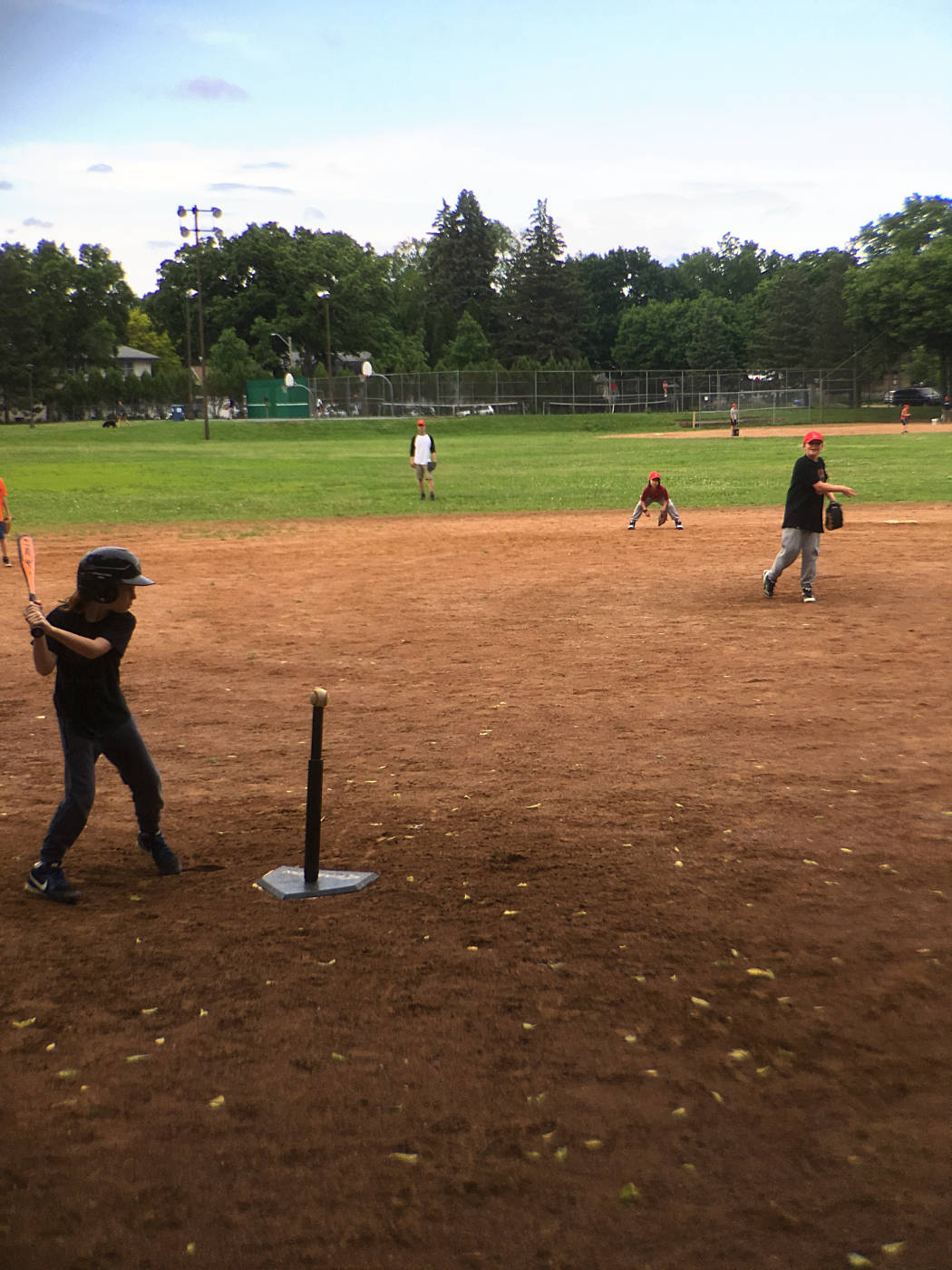 young person at bat with a baseball on a pole seen from behind homeplate looking out at other kids on the diamond