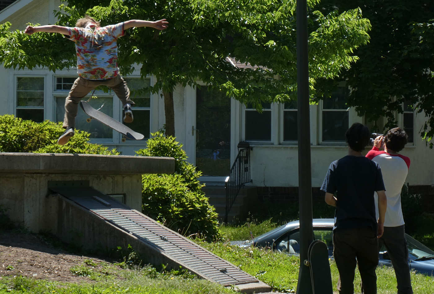 A skateboarding young man is in midair, arms outstretched, skateboard flipped sideways underfoot, above a narrow cement ramp down to the ground, as two companions watch, and one is holding a video camera