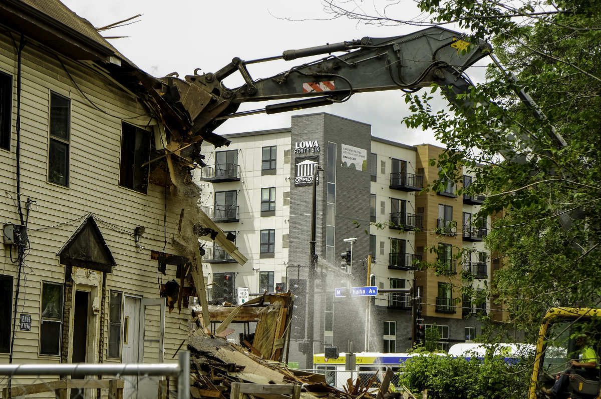A construction excavator tears into an aged two-story yellow clapboard building, with a new five-story building in background