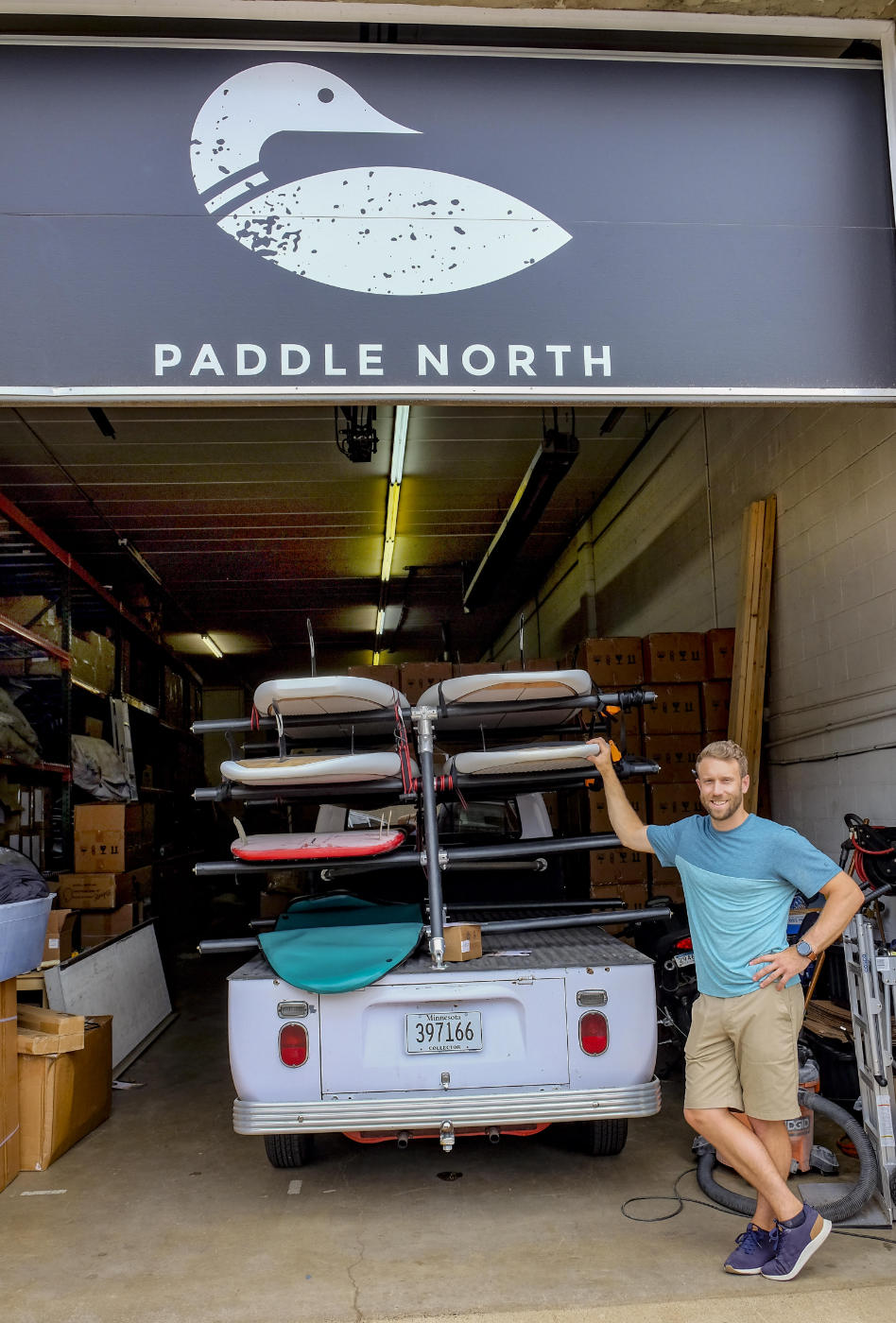 man in T-shirt and shorts leaning against back of a small truck carrying six paddleboards on a rack, in a garage port with a painted sign on top with PADDLE NORTH and loon logo