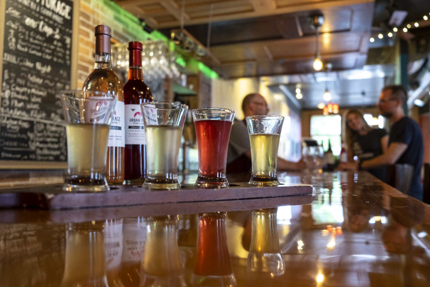 Four glasses of cider colored pale straw to light red with bottles and background out of focus with bar and people