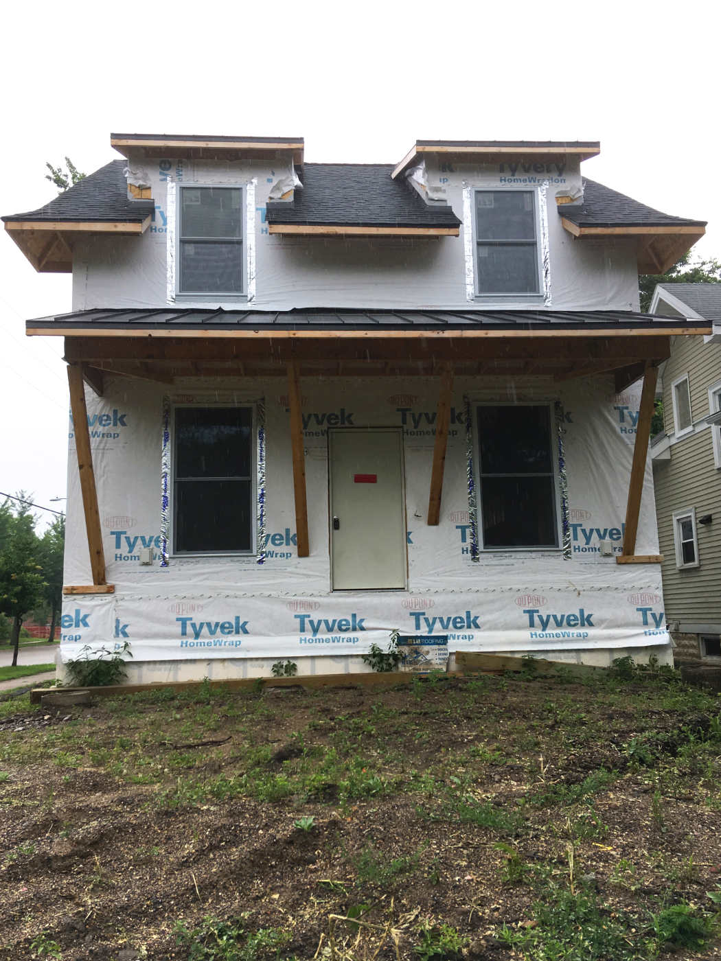 two-story house under construction with Tyvek wrap on walls with silver foil trim on new windows on dirt lot with scrub weeds