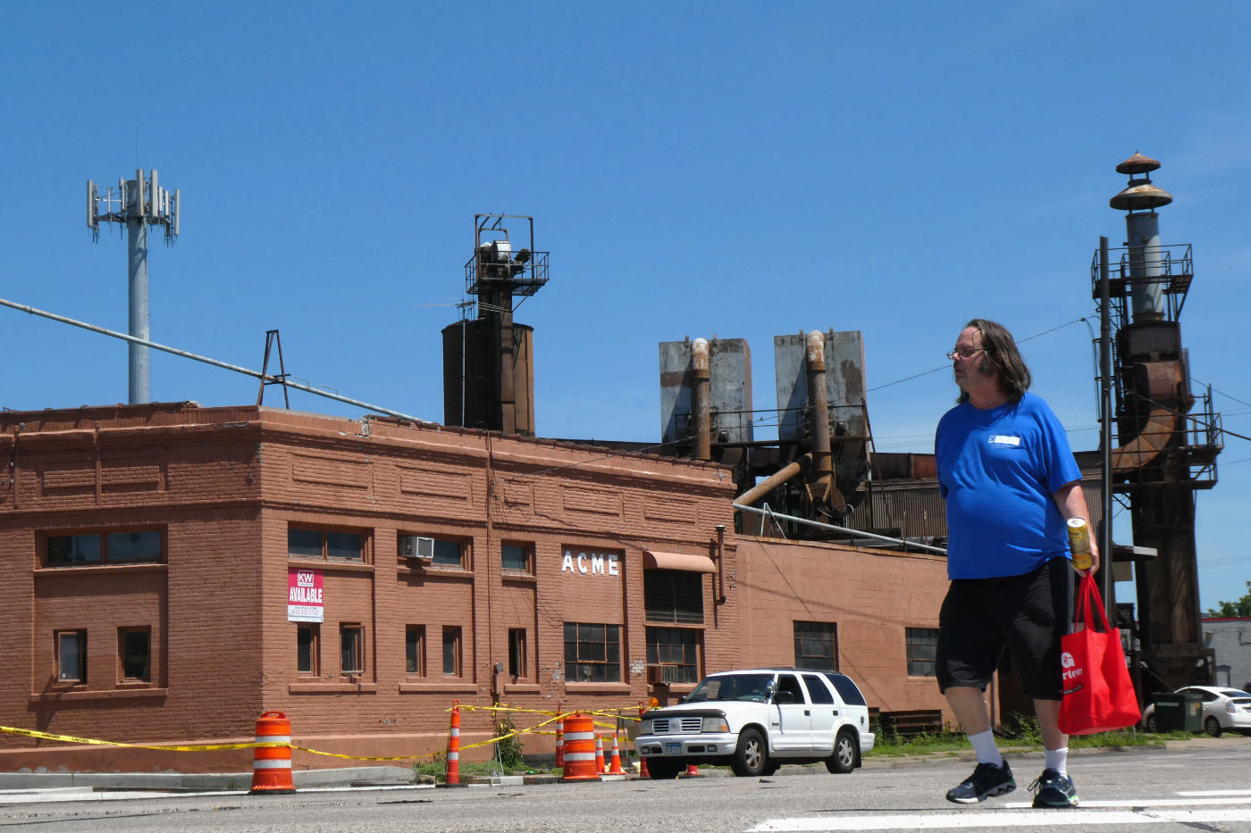 man in shorts with red shopping bag crossing street with ACME industrial building in background and bright blue sky