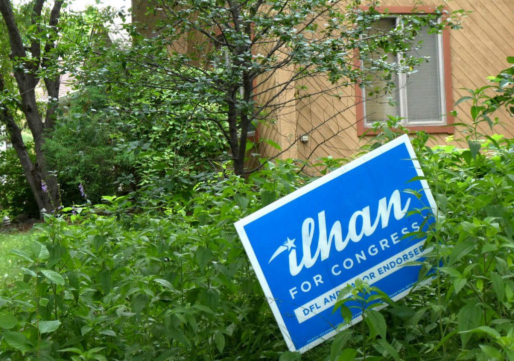 Ilhan for Congress blue and white lawn sign amidst green foilage with tan house and window in background