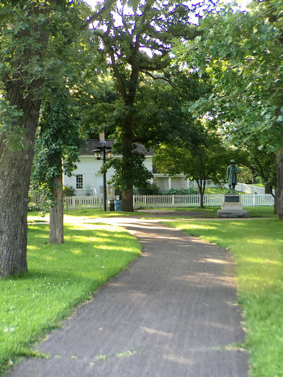 gray sidewalk leading up to a white house and picket fence with a statue of a man in the yard