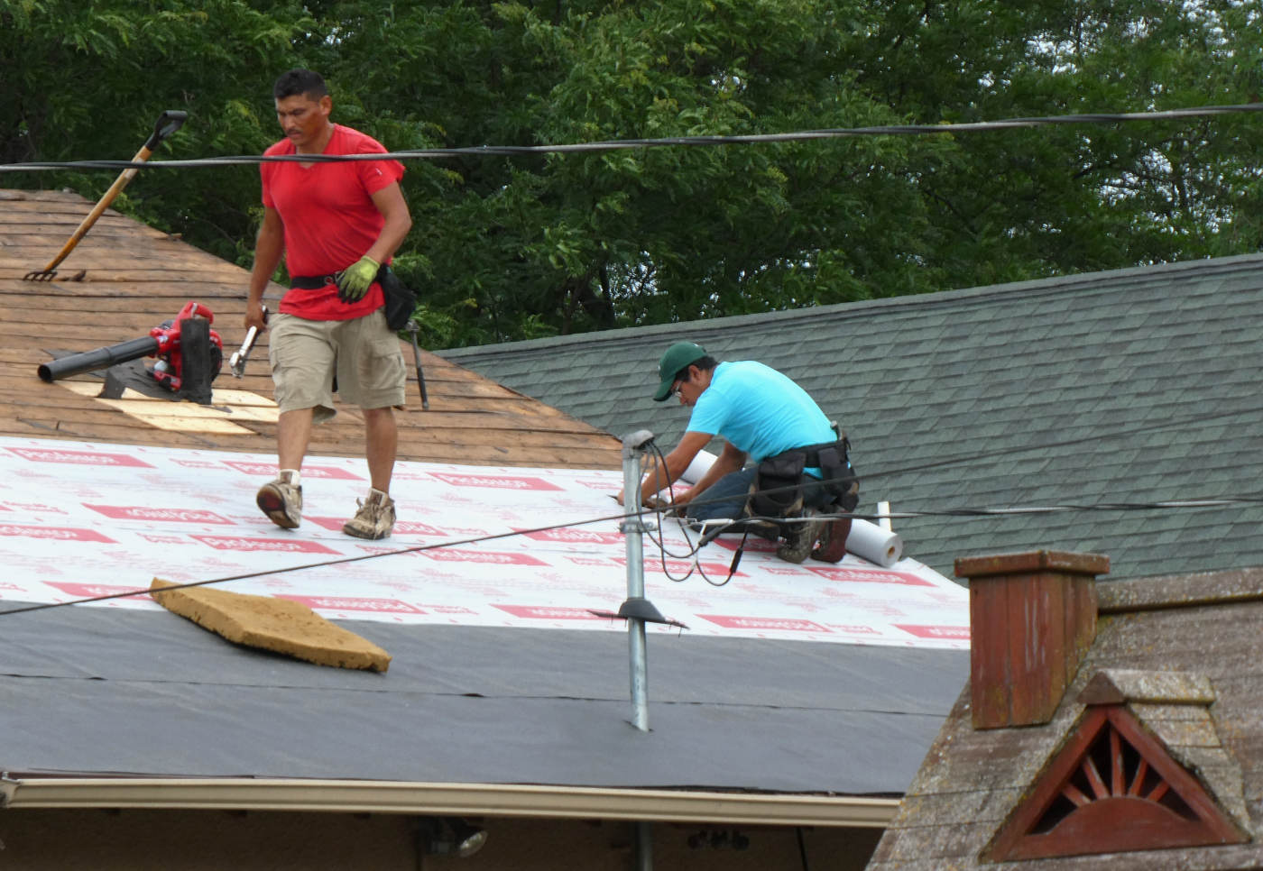 atop a roofing job, a man in a red shirt walking and a man in a blue shirt kneeling and hammering