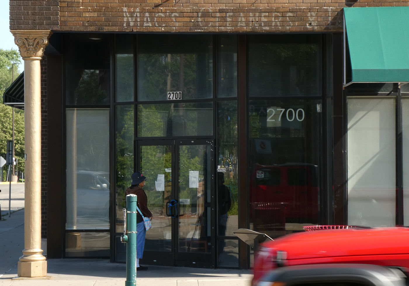 Person at the door of a closed storefront with a faded sign painted overhead on bricks reading MAC'S CLEANERS & ...