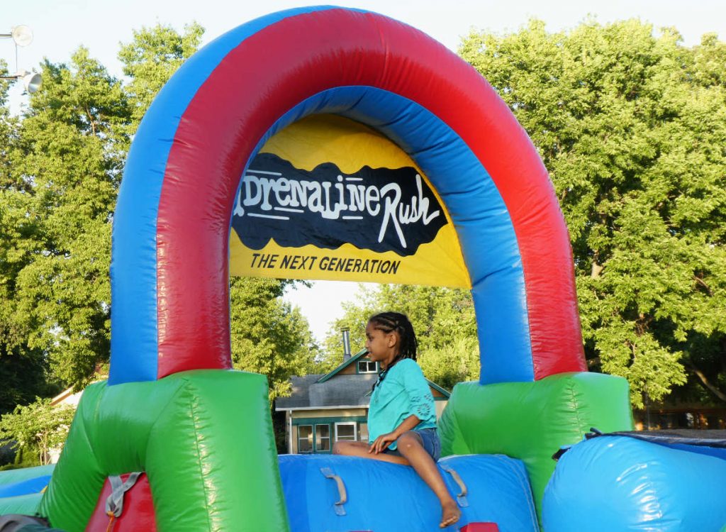young woman sitting atop an inflatable kids play course with an arch overhead and sign reading Adrenaline Rush: The Next Generation 
