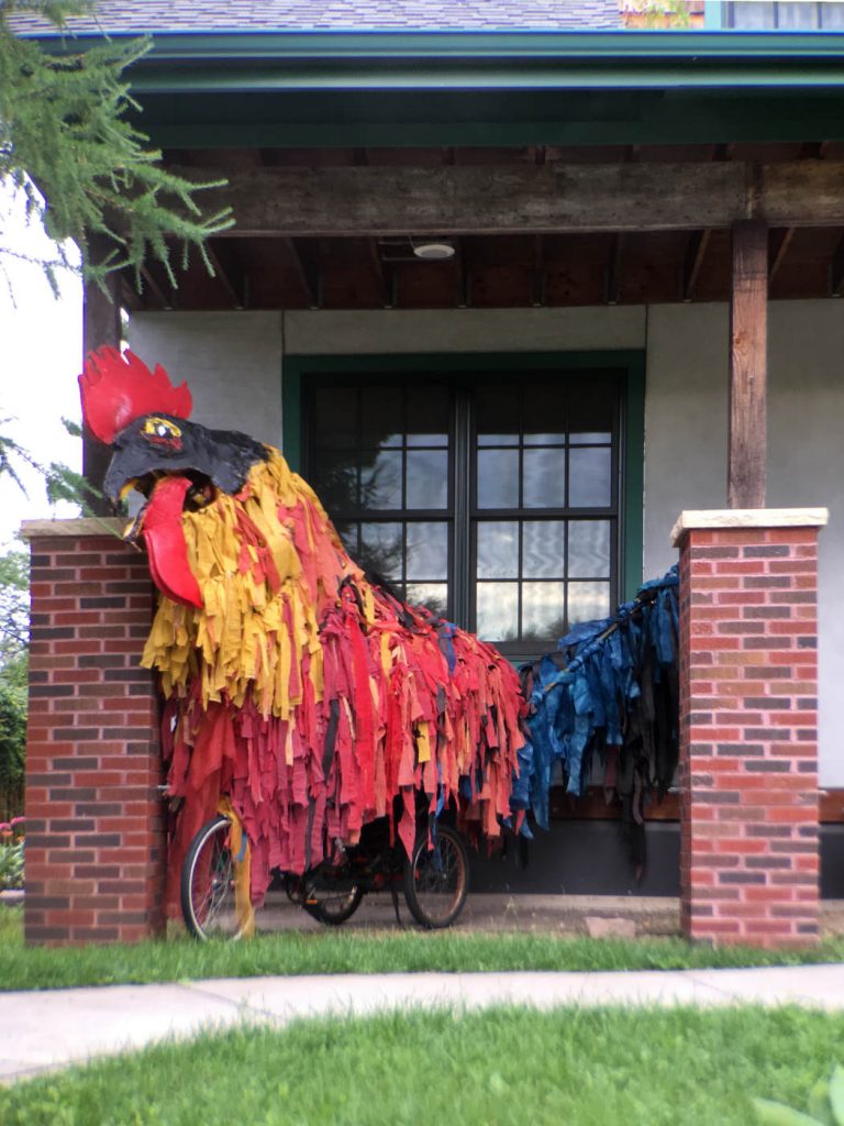 large paper papier-mâché and crepe paper rooster atop a bicycle on  a house porch