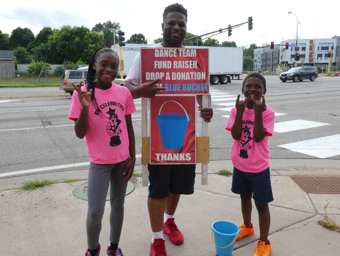 two young girls and a man posing for camera with a sign reading: DANCE TEAM FUNDRAISER, DROP A DONATION with picture of a blue bucket, and a blue bucket on the ground