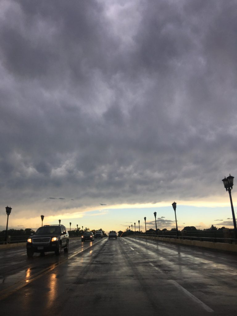 cars with headlights shining on wet pavement with dark gray storm clouds and blue to yellow sky on horizon