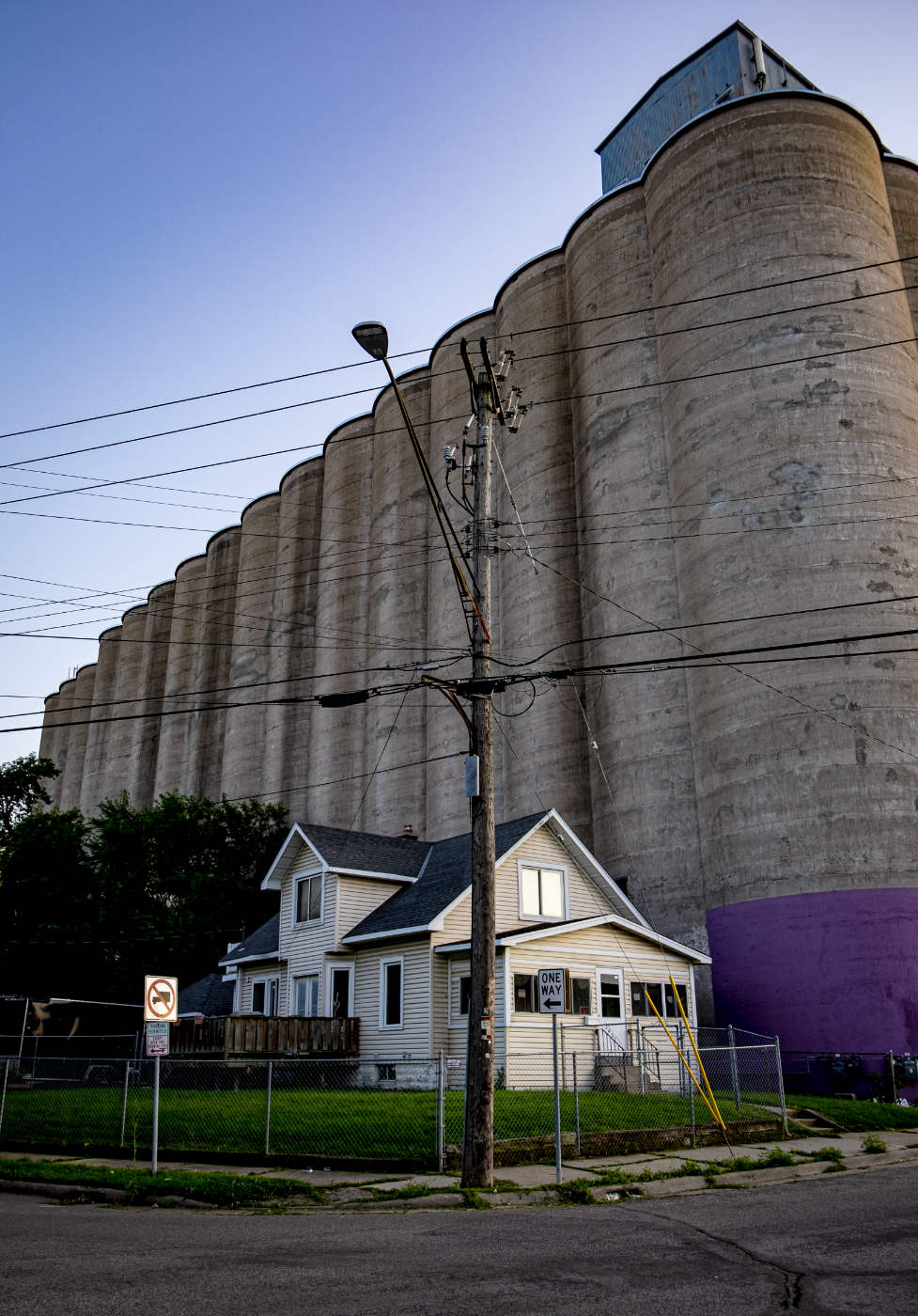 Tall gray grain elevators receding against a blue sky towering over a bungalow house in front