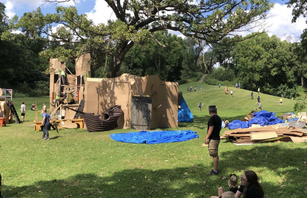kids play around a large cardboard castle in a green field surround by hills and trees in the background