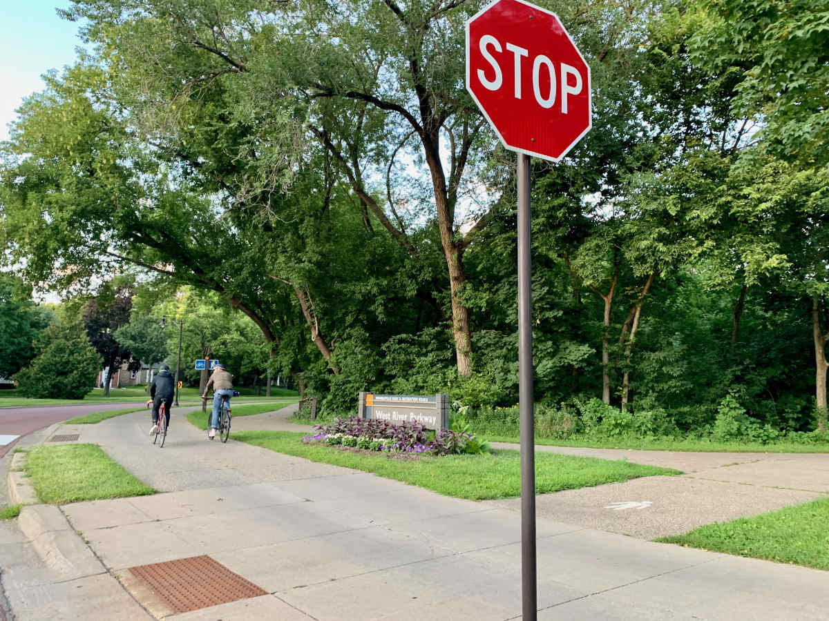 two people ride bicycles on a bike path past a wooden sign reading "WEST RIVER PARKWAY" along with a Stop sign in the foreground