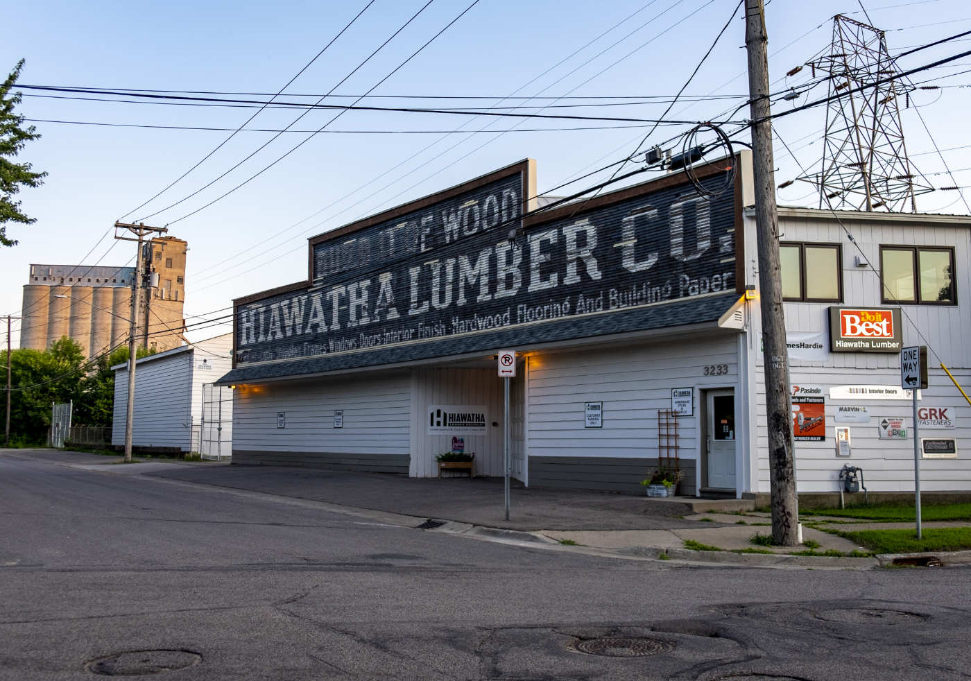 Long single-story white industrial building with large white on dark gray signage across the top reading: HIAWATHA LUMBER CO. with other faded lettering