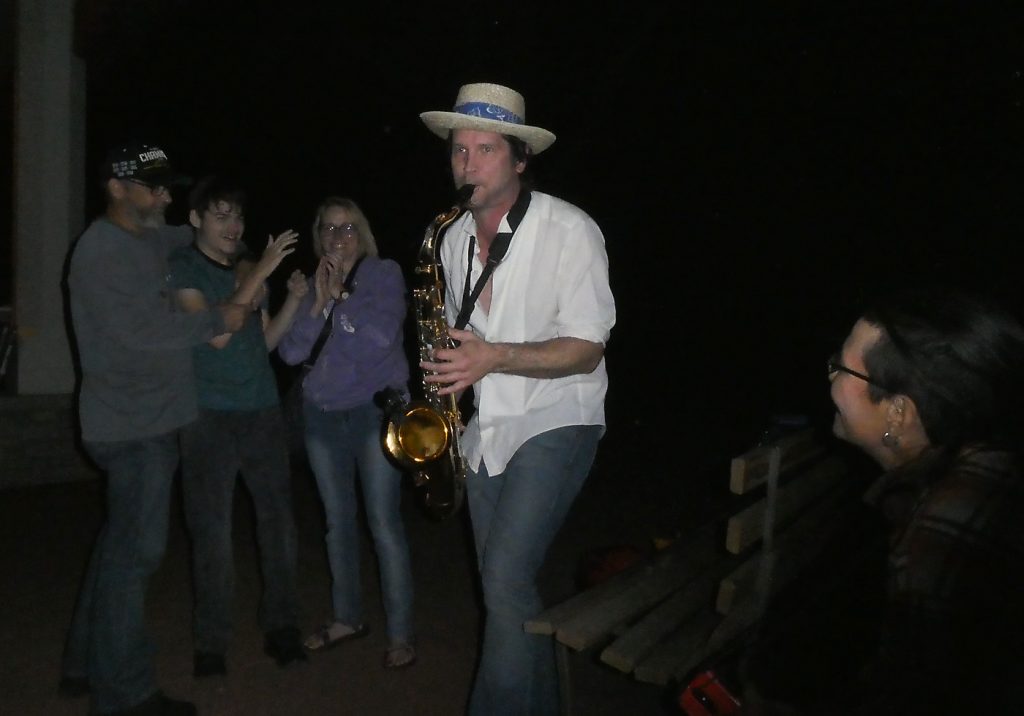 saxophone player in white shirt, jeans, and straw hat playing in the dark as a woman looks on in the foreground and three people clap hands along in the background