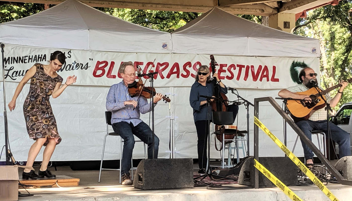 On stage is a woman clogging, a man fiddling, a woman on bass, and a man on guitar, with a banner in the background for Minnehaha Falls "Laughing Waters" Bluegrass Festival