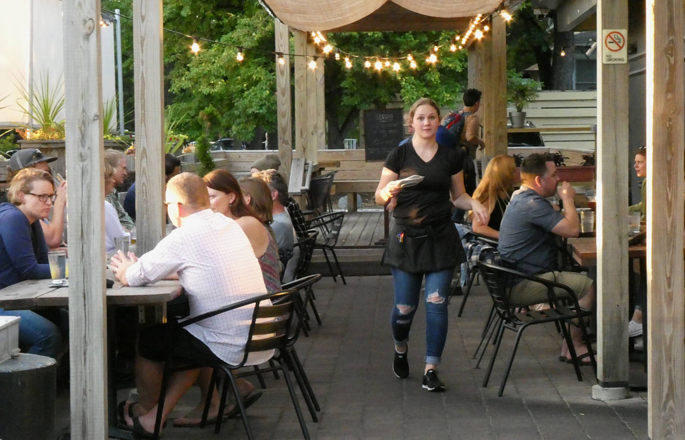 waitress walking forwards between cafe tables with diners, lights overhanging an outdoor patio area with wooden beams surrounding