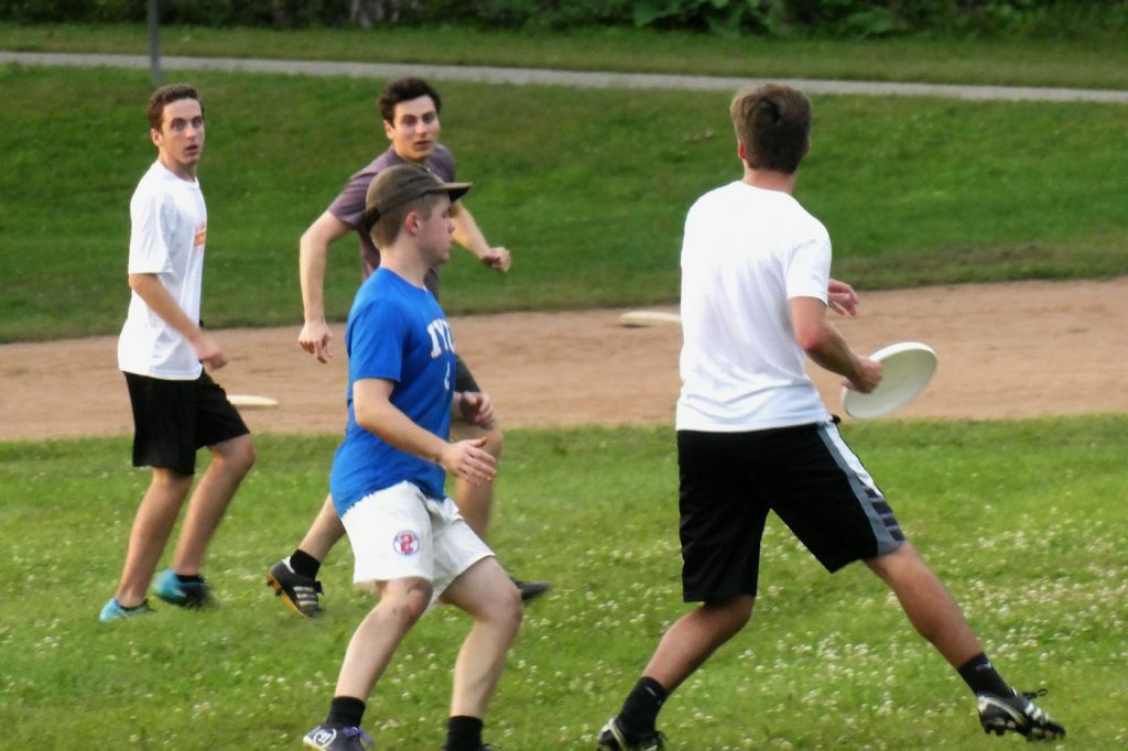 One young man in T-shirt and shorts runs with a frisbee ready to throw while three other players run to follow