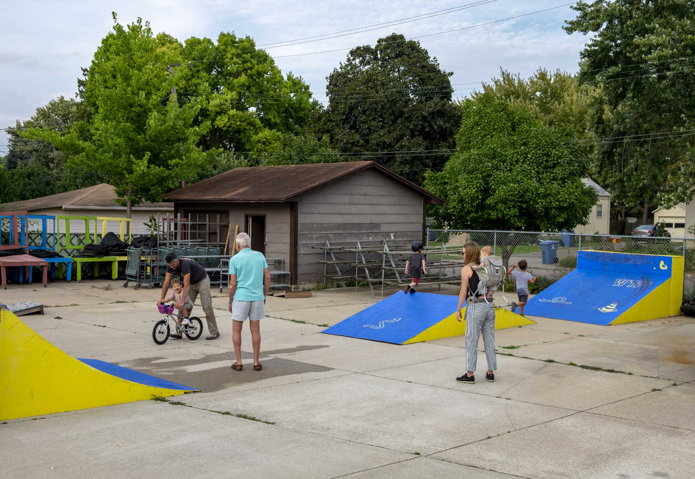 a few adults play with a few youngsters, one on a small bicycle, in a paved lot with small ramps