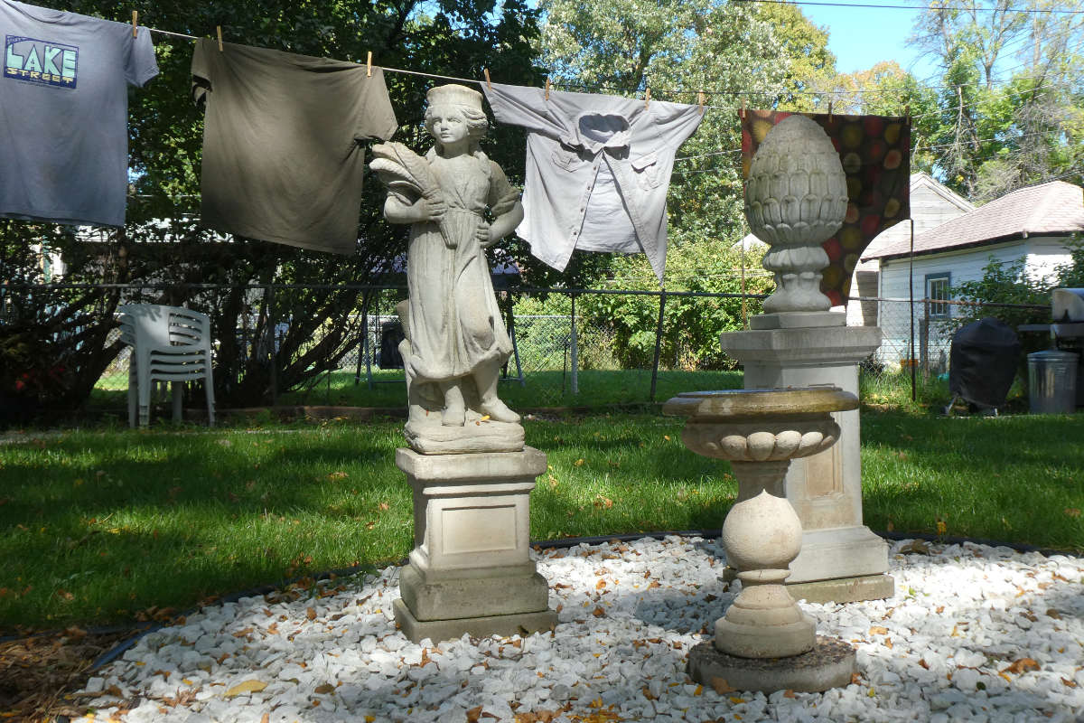 Statue of girl with wheat shaft and statue of finial on crushed stone in front of a laundry line in a backyard