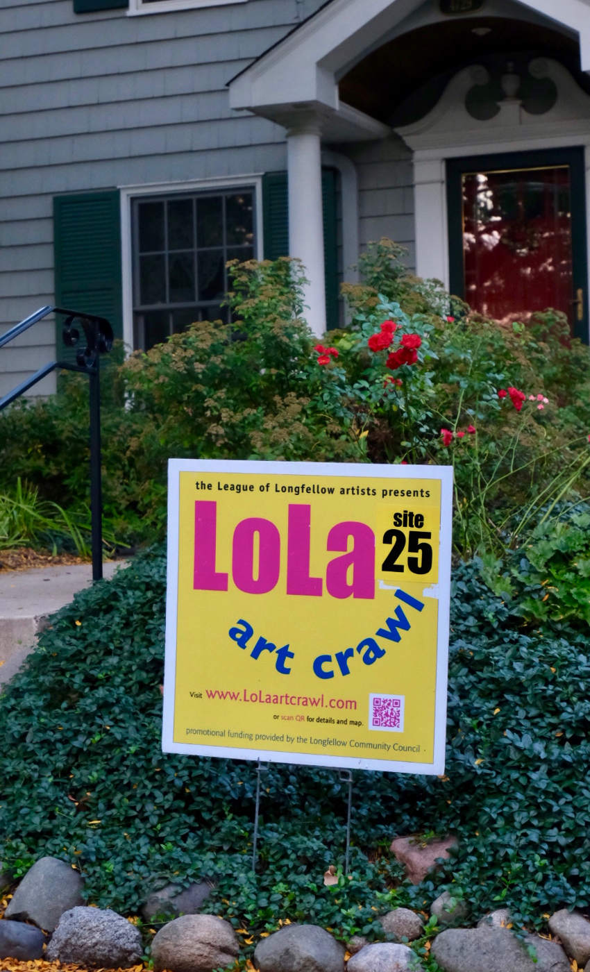 Yellow yard sign with pink and blue lettering reading LoLa art crawl, and with rosebushes and a house front door in background